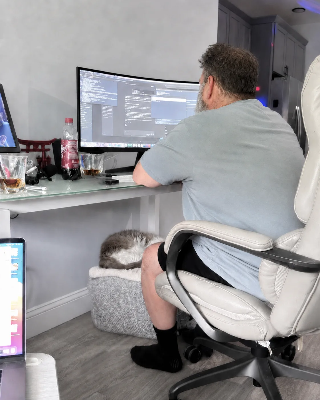 Greg coding the Chiro Connect system at his desk, two monitors of code in front of him, a bourbon and the office cat nearby.