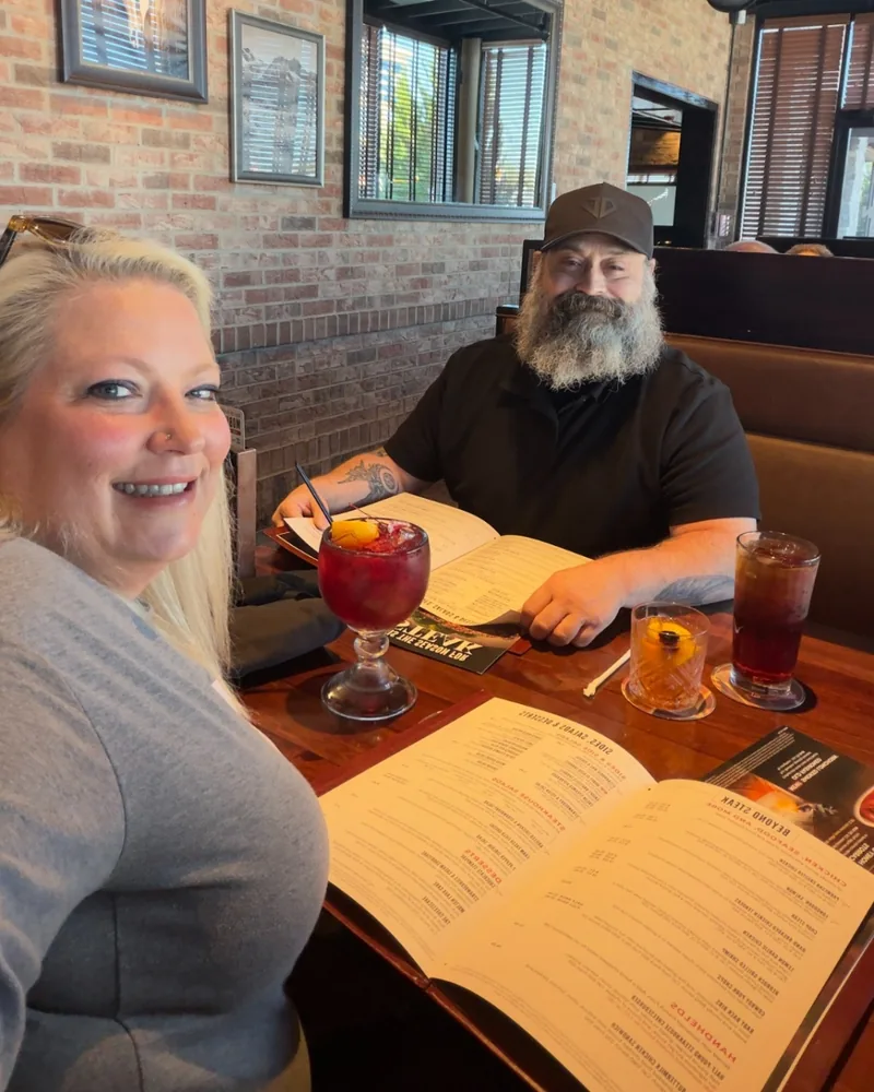Nerissa smiling in the foreground of a restaurant booth while Greg, in a cap and black shirt, reads the menu across the table with an old fashioned in front of him.