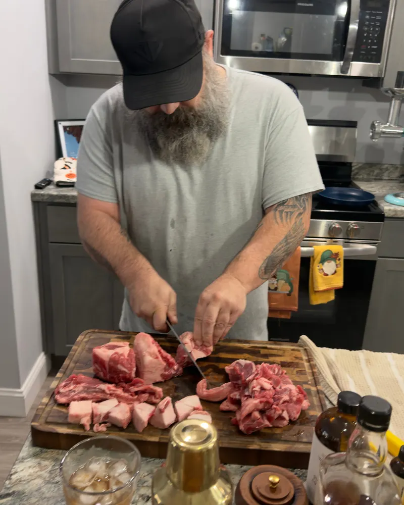 Greg, in a black cap and gray t-shirt, cutting raw beef into chunks on a wooden board at the kitchen counter, with a meat grinder on the counter behind him.