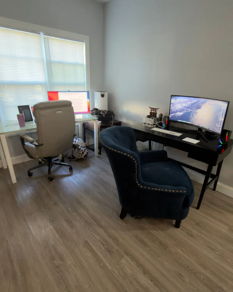 The new home office: a white desk by the window with a gray chair, a black desk with a monitor on the far wall, and a navy blue armchair sitting in the middle of the room.