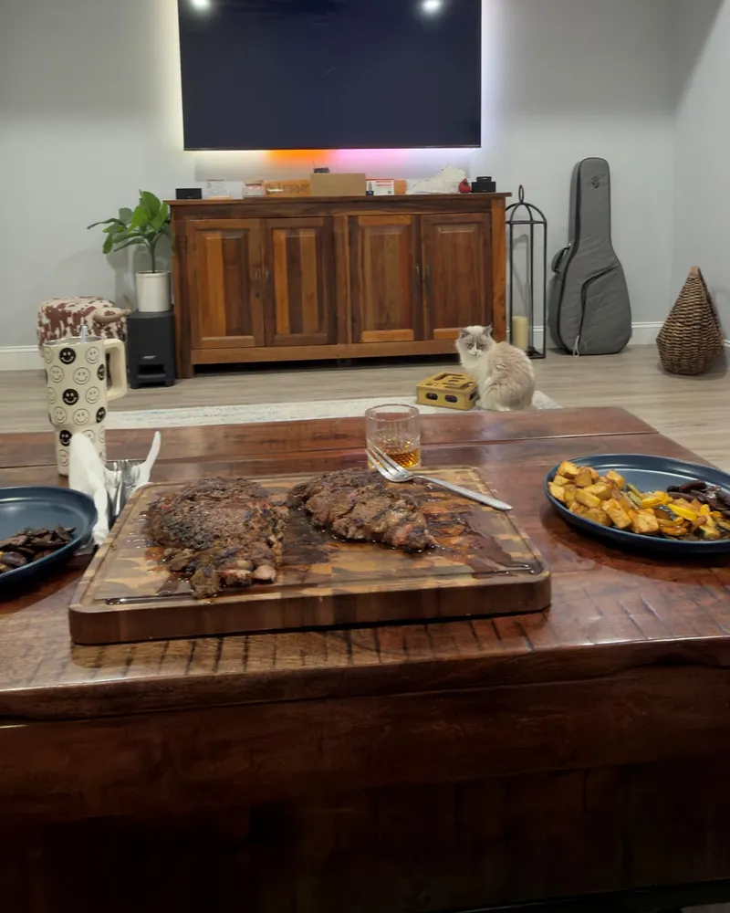 Two ribeyes on a cutting board and a glass of bourbon, set up in front of the TV.