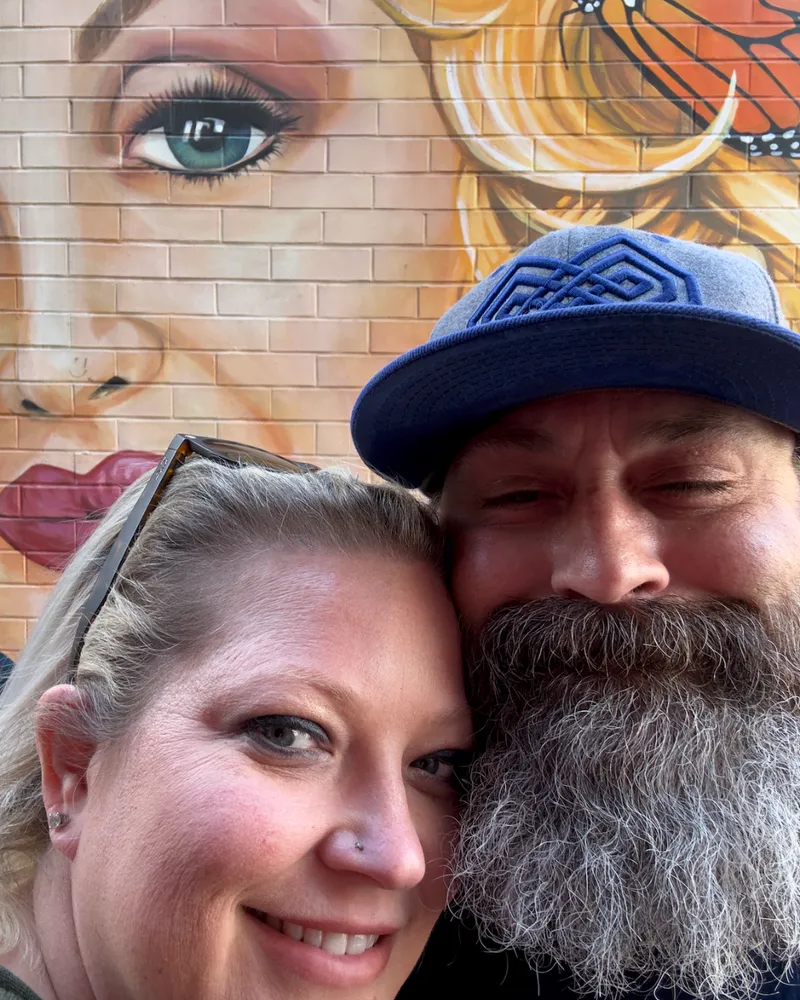Nerissa and Greg standing together beneath a large mural of Dolly Parton painted on a brick wall.