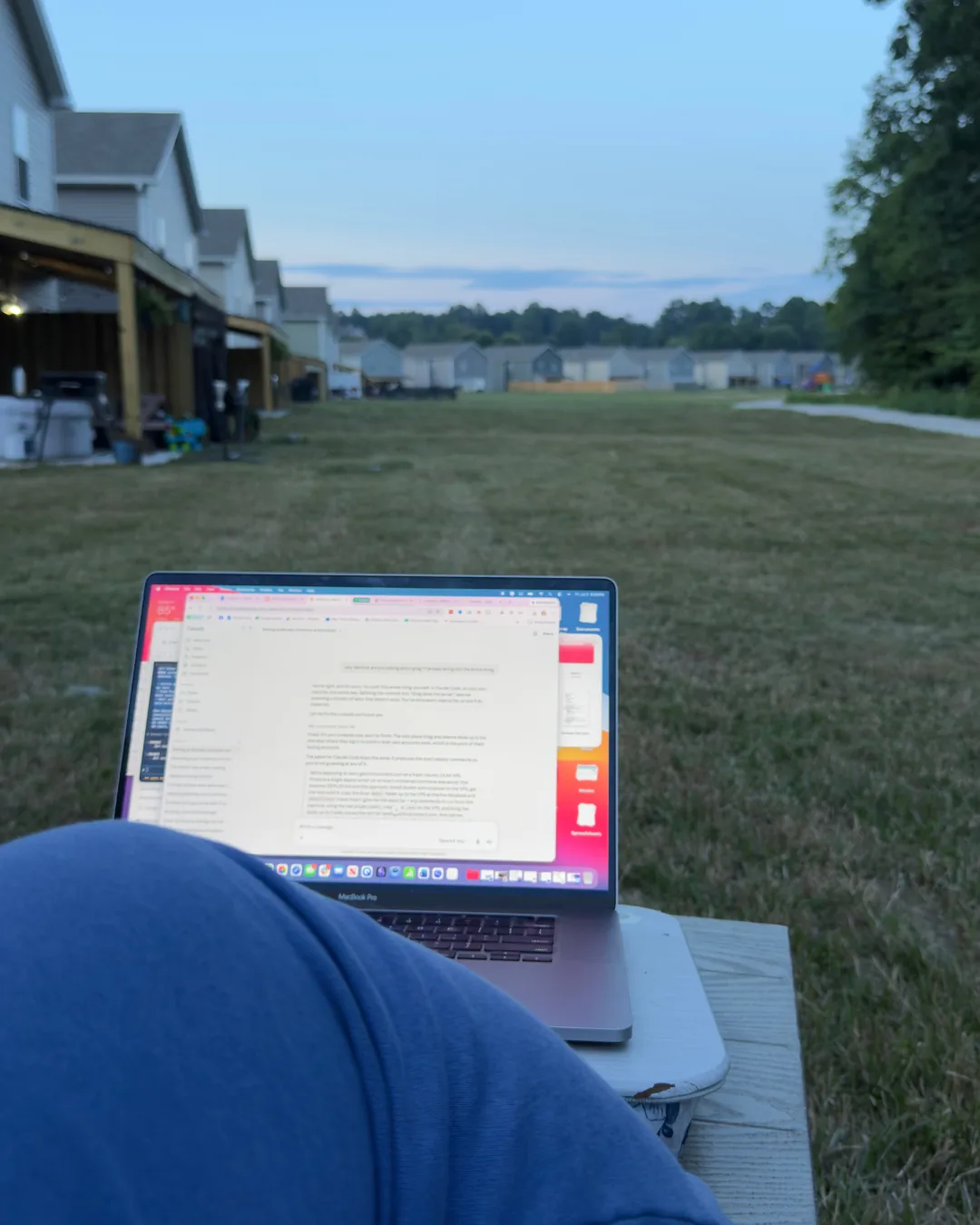 Nerissa's legs in blue sweatpants with a MacBook open to Claude on a bench, houses and grass behind her at dusk on the Fourth of July.
