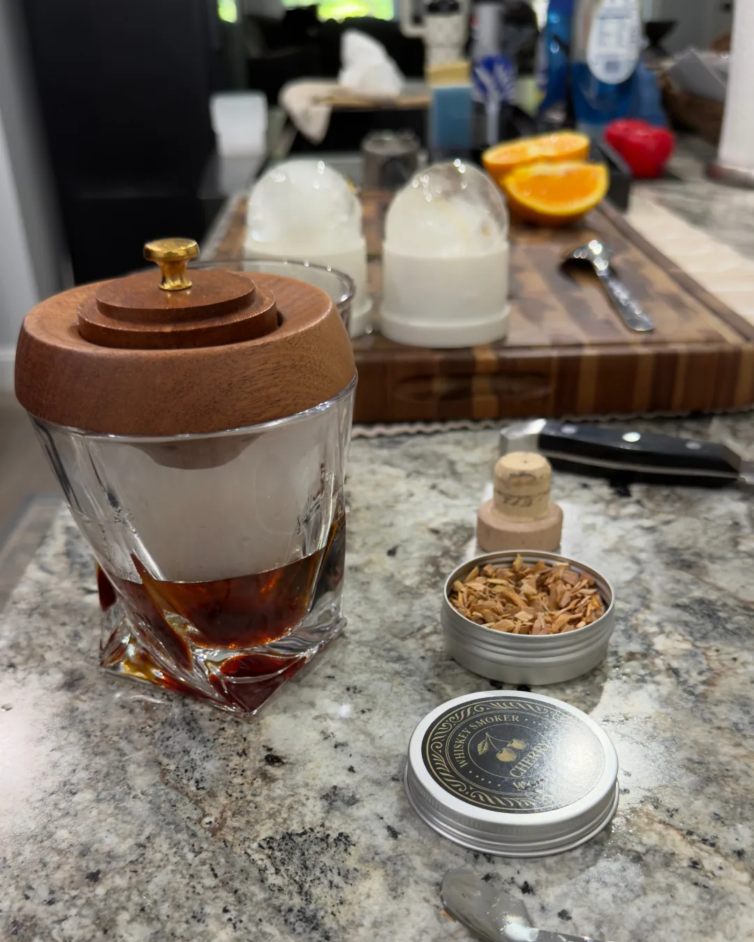 Greg's Old Fashioned station on the counter: a whiskey glass with a wooden smoker lid, ice balls, orange halves, cork, and a tin of cherry wood chips.