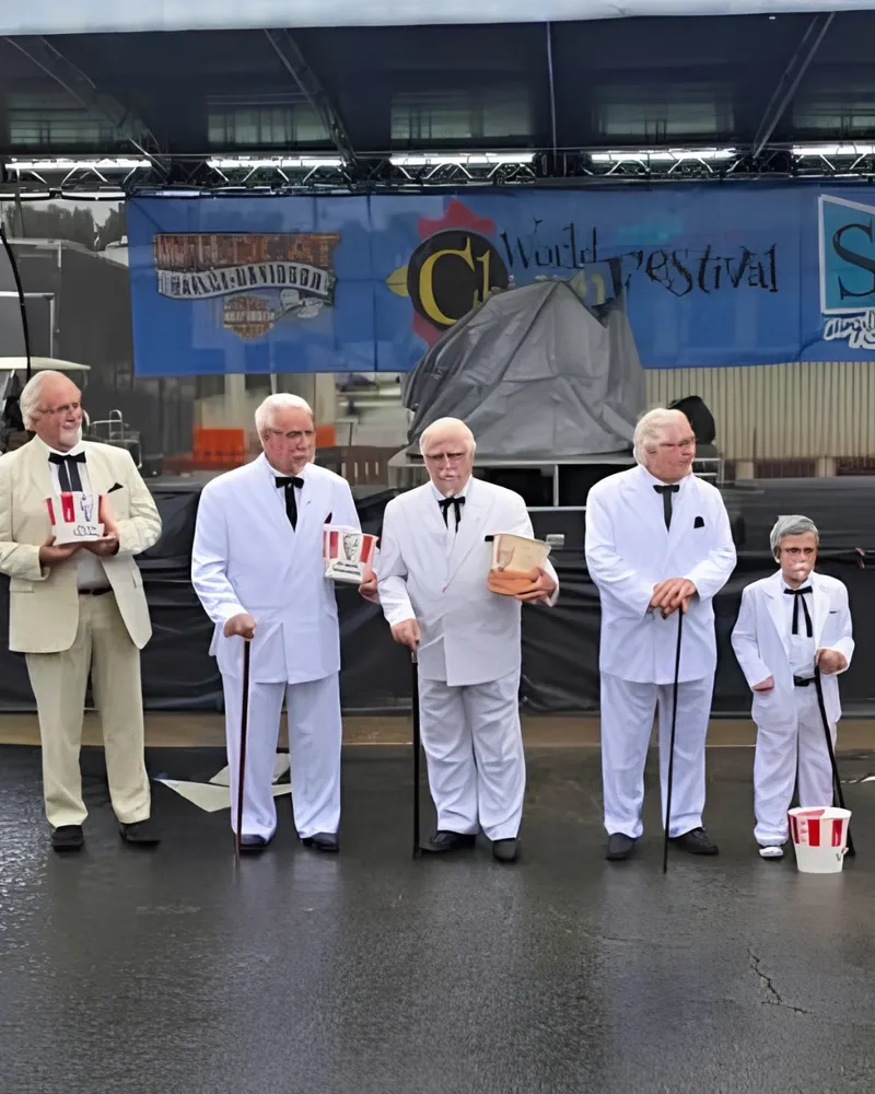 Five Colonel Sanders look-alikes in white suits with canes and chicken buckets lined up in front of the World Chicken Festival stage.