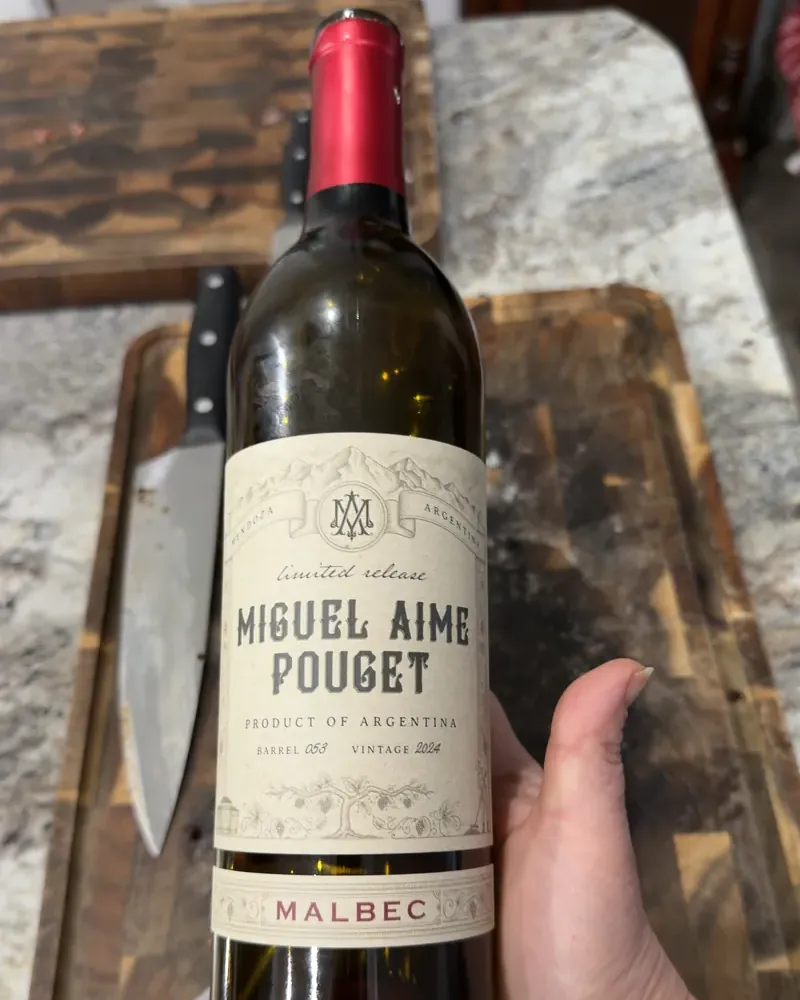 A hand holding a bottle of Miguel Aime Pouget Malbec over a wooden cutting board on a granite counter, with a large kitchen knife lying beside it.