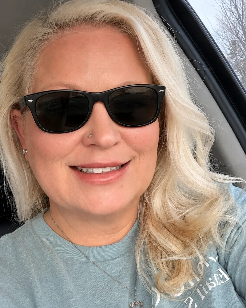 Nerissa taking a car selfie in black sunglasses and a light blue sweatshirt, smiling.